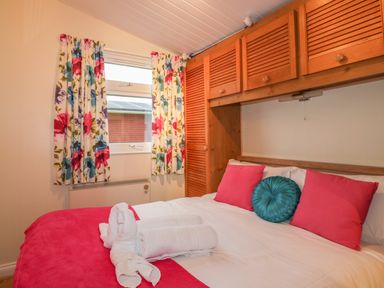 A bedroom with a bed with pink and blue pillows white towels floral curtains and wooden cupboards at Tiggy Winkle in St Merryn near Padstow