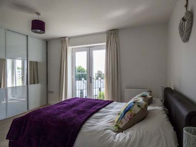 A bedroom with a bed covered by a purple blanket and floral pillows next to glass doors and a wardrobe with sliding doors at Tehidy Charlestown