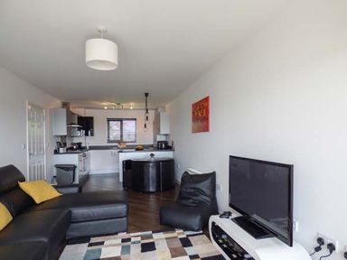 An open living room and kitchen with a black sectional sofa with yellow pillows a patterned rug a flat screen tv on a white stand and a round black dining table at Tehidy in Charlestown