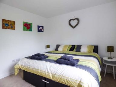 A bedroom with a double bed featuring striped bedding and folded towels on top with a side table and lamps and paintings on the wall at Tehidy in Charlestown