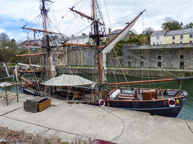 A docked sailing ship with masts and rigging by a stone wall and buildings at Tehidy in Charlestown