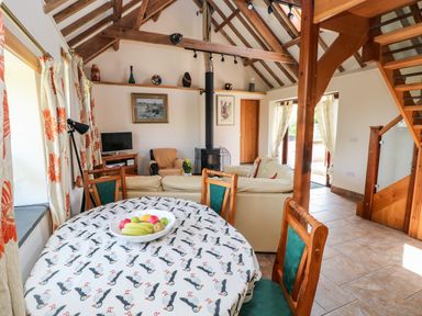 A living room with a dining table covered with a patterned tablecloth a bowl of fruit sofas a wood burning stove wooden staircase and patterned curtains at The Long Barn in Hasguard near Little Haven