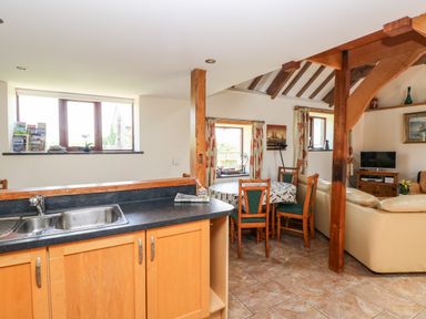 A kitchen area with a double sink and wooden cabinets overlooking a dining area with a round table and green cushioned chairs beside a living room with a beige sofa and a television at The Long Barn in Hasguard near Little Haven