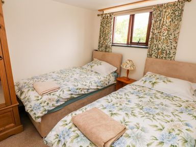 A bedroom with two single beds with floral bedding a wooden nightstand with a lamp a window with patterned curtains and a wooden wardrobe at The Long Barn in Hasguard near Little Haven