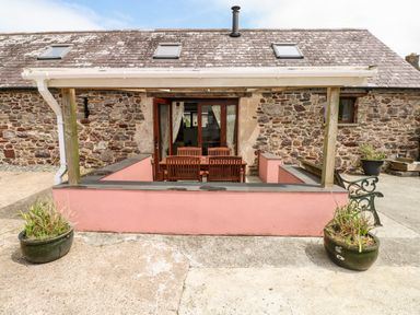 A stone building with a small covered outdoor seating area with wooden table and chairs and two large planters at The Long Barn in Hasguard near Little Haven