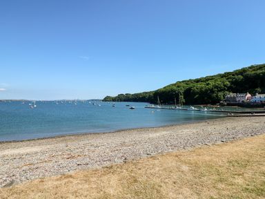 A pebble beach with grassy area and boats on water near houses by trees at The Long Barn in Hasguard near Little Haven