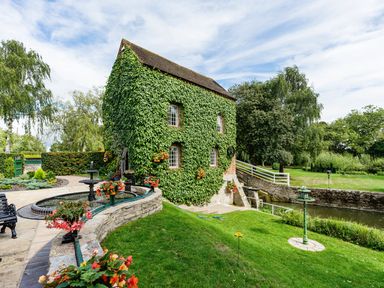 An ivy-covered stone building next to a circular fountain and a grassy area with trees and a small canal at Grafton Mill Grafton Flyford near Pershore