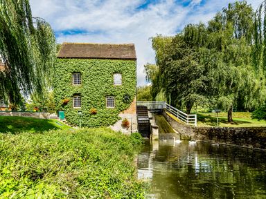 A watermill with ivy-covered walls beside a river with swans and surrounding trees at Grafton Mill in Grafton Flyford near Pershore