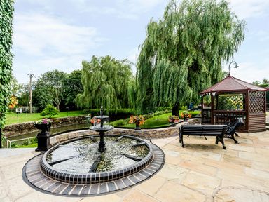 A garden patio with a circular water fountain benches and a wooden gazebo near large trees at Grafton Mill in Grafton Flyford near Pershore