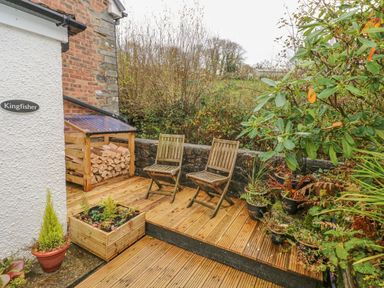 An outdoor wooden deck with two wooden chairs firewood storage and potted plants at Kingfisher in Blaenwaun