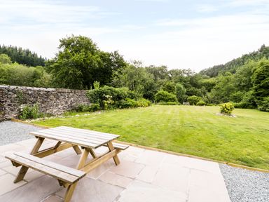 A wooden picnic table on a stone patio overlooking a grassy lawn with trees and a stone wall at Hen Gelligemlyn - The Annexe in Ganllwyd