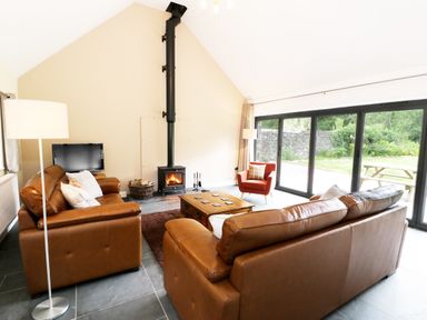 A living room with brown leather sofas a wood stove and large windows at Hen Gelligemlyn - The Annexe in Ganllwyd