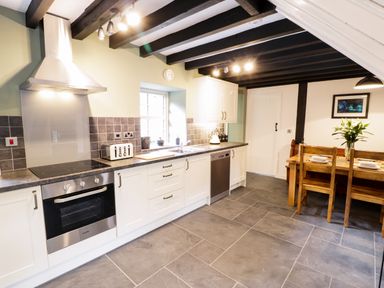 A kitchen with white cabinets a stove a toaster a kettle a sink a dining table with chairs and flowers at Hen Gelligemlyn - The Annexe in Ganllwyd