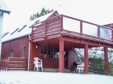 A red wooden house with a snow-covered roof and balcony with plastic chairs on the ground at Strathavon Chalet in Aviemore