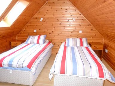 A bedroom with two single beds with striped bedding and wooden nightstands under a slanted wooden ceiling at Strathavon Chalet in Aviemore