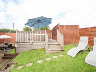 A fenced backyard with a wooden deck stairway leading to a patio with an umbrella and a hot tub two lounge chairs on the grass and a small table with an umbrella and chairs at Bwthyn Ty Newydd in Llangadfan