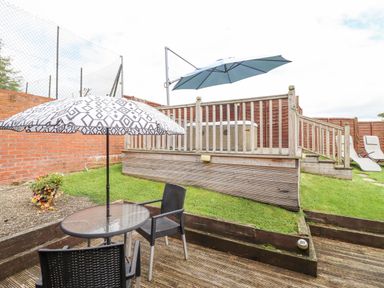 An outdoor patio area with a glass table, two black chairs, patterned umbrella, wooden deck, raised hot tub with blue umbrella, grass, and two lounge chairs at Bwthyn Ty Newydd in Llangadfan
