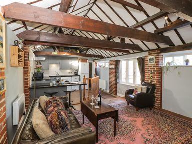 An open living room and kitchen area with exposed wooden beams a sofa and armchair a wooden coffee table and a patterned rug at Old Cart Shed Lyneham near Royal Wootton Bassett