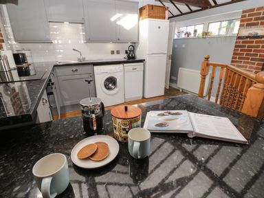 A kitchen with grey cabinets a washing machine a dishwasher and a fridge freezer with mugs a biscuit jar and a recipe book on the countertop at Old Cart Shed in Lyneham near Royal Wootton Bassett