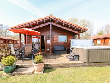 A wooden lodge with a deck featuring a hot tub table with chairs and an orange umbrella at Bittern Lodge in Tattershall Lakes Country Park