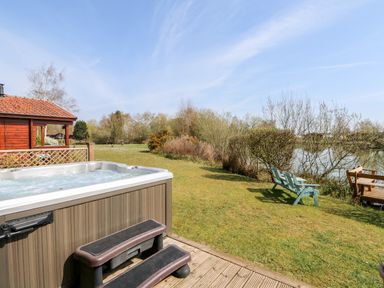 A hot tub with steps on a deck near a grassy area with chairs and a lake at Bittern Lodge in Tattershall Lakes Country Park