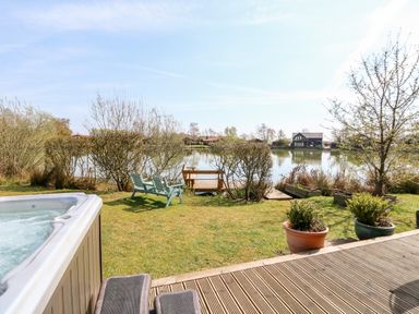 A garden with lawn chairs near a wooden dock by a lake and a hot tub on a wooden deck at Bittern Lodge in Tattershall Lakes Country Park