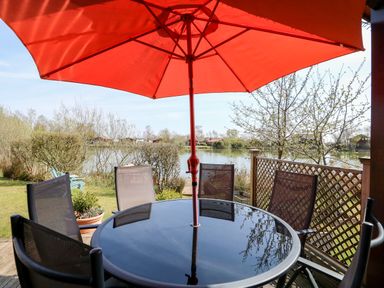 Outdoor patio area with a round glass table and chairs under a red umbrella near a lake at Bittern Lodge in Tattershall Lakes Country Park