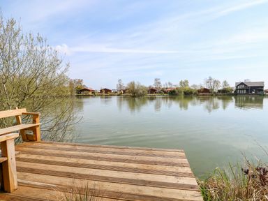 A wooden deck with a bench overlooking a lake with houses and trees across the water at Bittern Lodge in Tattershall Lakes Country Park