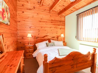 A bedroom with wooden bed frame bedside tables lamps dresser and a window with blinds at Bittern Lodge in Tattershall Lakes Country Park