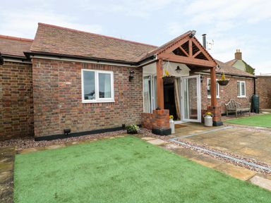 A single-story brick house with a tiled roof an open glass door a wooden porch hanging flower baskets green artificial turf and a paved patio area at The Stable in Berkeley