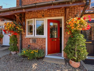 A front entrance with a red door surrounded by hanging flower baskets and potted plants at Camerons Cottage in Swaffham
