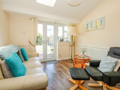 A sitting room with cream sofa with cushions black leather armchair and glass doors leading to a garden at Camerons Cottage in Swaffham
