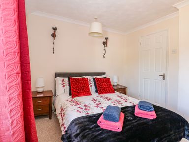 A bedroom with a double bed with floral bedding red pillows folded towels bedside tables lamps and closed door at Camerons Cottage in Swaffham