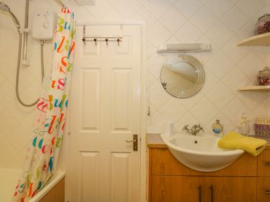 A bathroom with a shower curtain, white door, round mirror, sink, wooden cabinet, and shelves with jars at Camerons Cottage in Swaffham