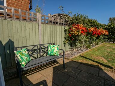 A garden with a metal bench with two green leaf patterned cushions next to a painted wooden fence and flowerbed at Camerons Cottage in Swaffham