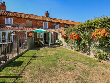 A backyard with a grass lawn a fence chairs table with umbrella and flowers on a fence at Camerons Cottage in Swaffham