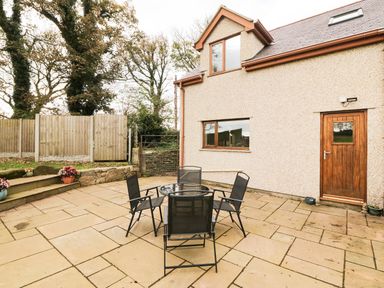 A patio area with a glass table and four black chairs outside a house with a wooden door and windows at Bodneithior in Llanerchymedd