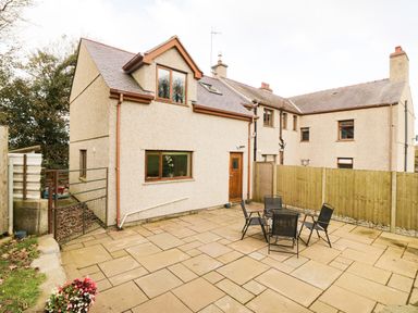 A paved patio area with a black table and four chairs next to a beige two-story house with brown window frames and a wooden door at Bodneithior in Llanerchymedd