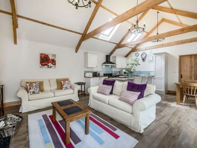 An open plan living room and kitchen with wooden beams on the ceiling and sofas with cushions at Warbrook in Bradworthy