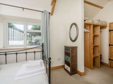 A bedroom with a metal bed frame and folded towels on the bed a window with curtains a small chest of drawers with an oval mirror above and an open wooden wardrobe at Warbrook in Bradworthy