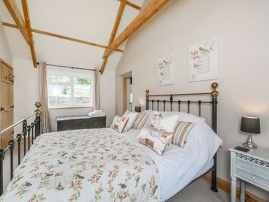 A bedroom with a bed featuring floral bedding and pillows wooden beams on the ceiling a window with curtains a wooden chest and a side table with a lamp at Warbrook in Bradworthy