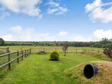 A grassy garden with a wooden picnic table small trees and a tunnel entrance surrounded by fencing at Warbrook in Bradworthy