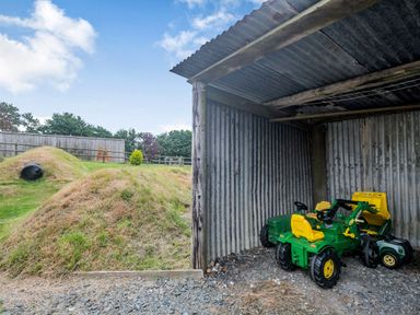 A corrugated metal shed with two green and yellow toy tractors next to a grassy area with hills and a tunnel at Warbrook in Bradworthy
