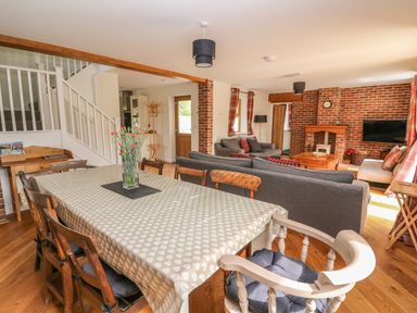 An open plan dining and living room with a dining table and chairs a vase with flowers a staircase sofas a brick fireplace and a television at The Farmhouse in Capel Bangor near Aberystwyth