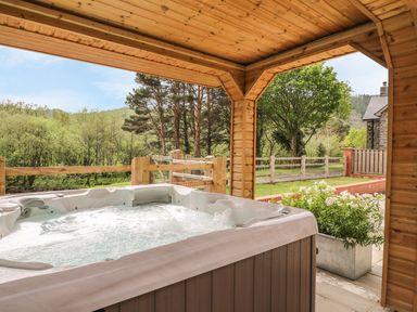 A covered outdoor patio with a hot tub and wooden fence overlooking trees and greenery at The Farmhouse in Capel Bangor near Aberystwyth