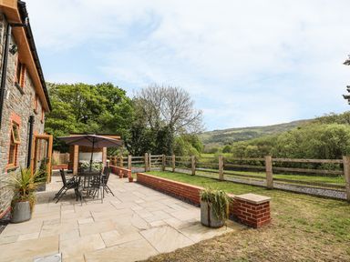 A patio with table chairs and umbrella next to a stone house with garden and hills in the background at The Farmhouse in Capel Bangor near Aberystwyth