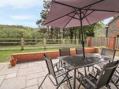 A patio with a glass table umbrella and six chairs overlooking a wooden fence and trees at The Farmhouse in Capel Bangor near Aberystwyth