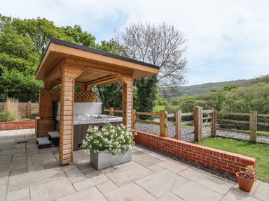A wooden gazebo with a hot tub on a stone patio next to a flower planter and a wooden fence with trees and hills in the background at The Farmhouse in Capel Bangor near Aberystwyth