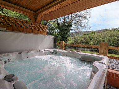 An outdoor hot tub with bubbling water under a wooden roof with a view of trees and hills at The Farmhouse in Capel Bangor near Aberystwyth