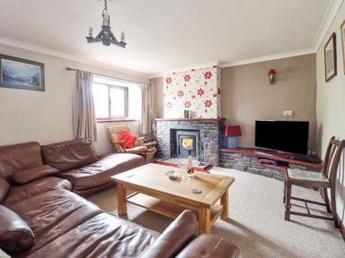 A living room with a brown leather sectional sofa a wooden coffee table a rocking chair a stone fireplace with floral wallpaper a TV on a stone shelf and a wooden chair at Haulfryn in Bethesda